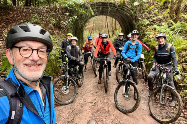 A group of muddy mountain bikers gather on a muddy track under an old single arch stone footbridge