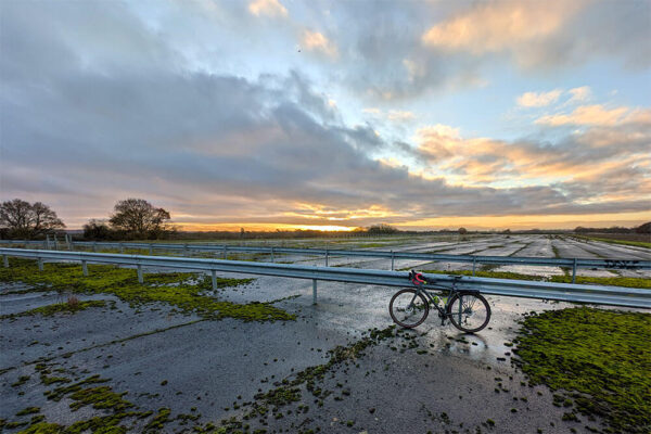 A Cannondale Topstone gravel bike rests against railings with the derelict Wisley airfield and a panoramic view of the setting sun low in the sky behind