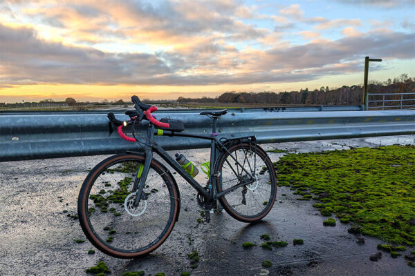 A Cannondale Topstone gravel bike rests against railings on the derelict Wisley airfield at sunset