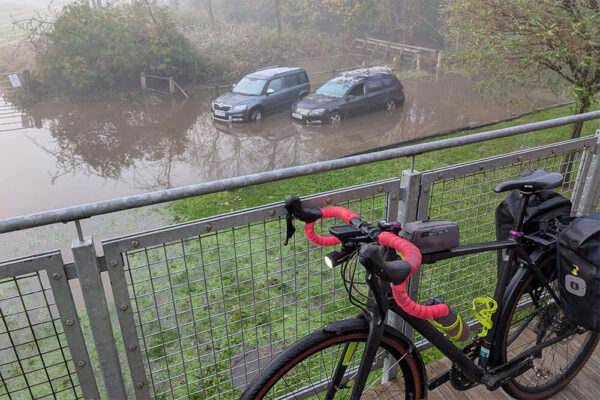 A gravel bike rests against railings above two cars on a road below, stranded and abadonned by flood waters behind